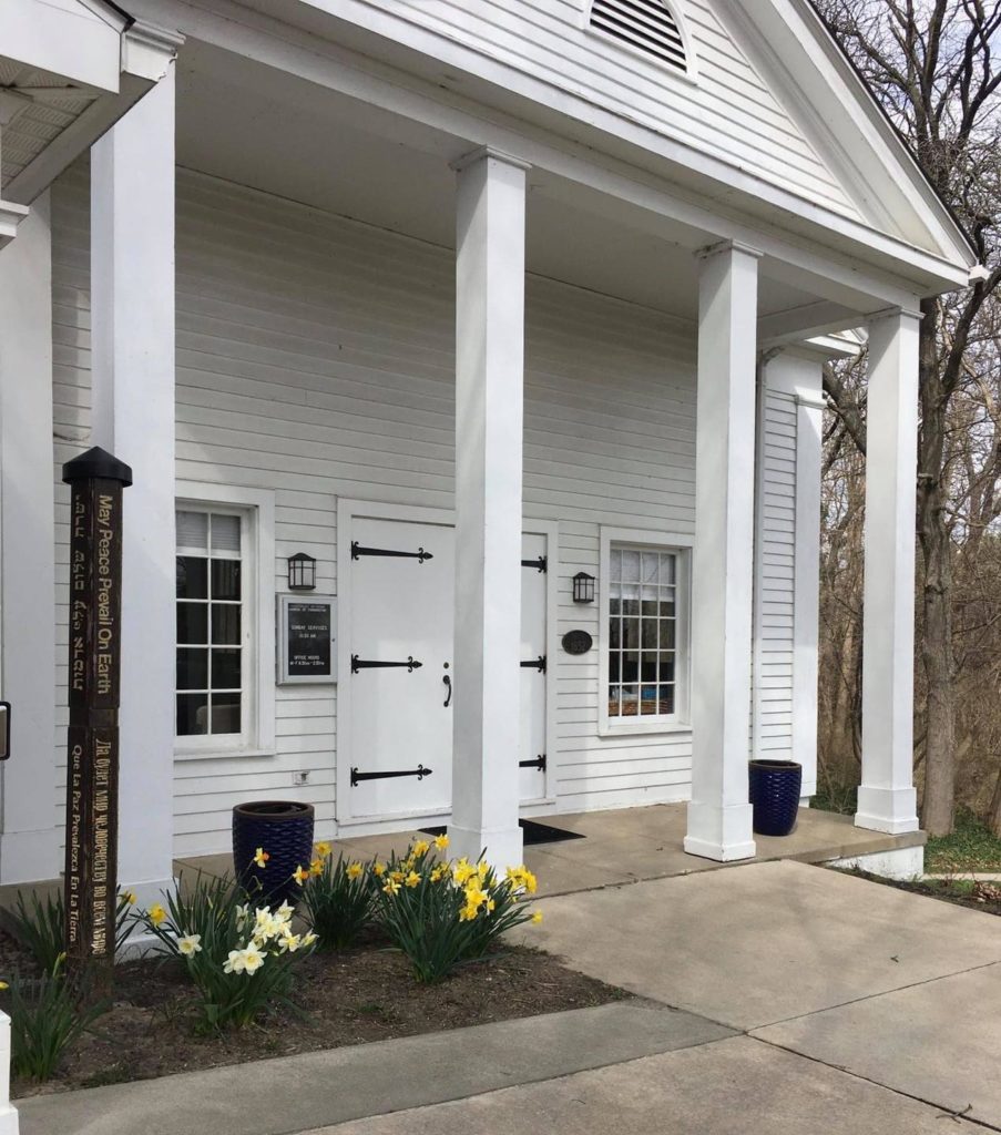 Side angle photo of UUCF's meeting hall front entrance. Four white pillars extend down, flanking a white door with black decorative hinges. Yellow flowers bloom in a garden to the left.