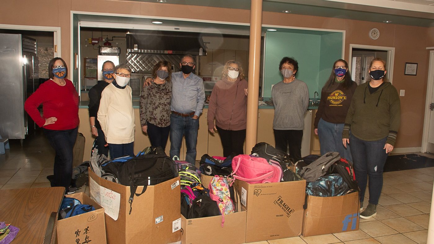 Nine UUCF social justice members pack backpacks for the homeless in 2020. Taken in UUCF dining hall, boxes filled with backpacks in front of the people.