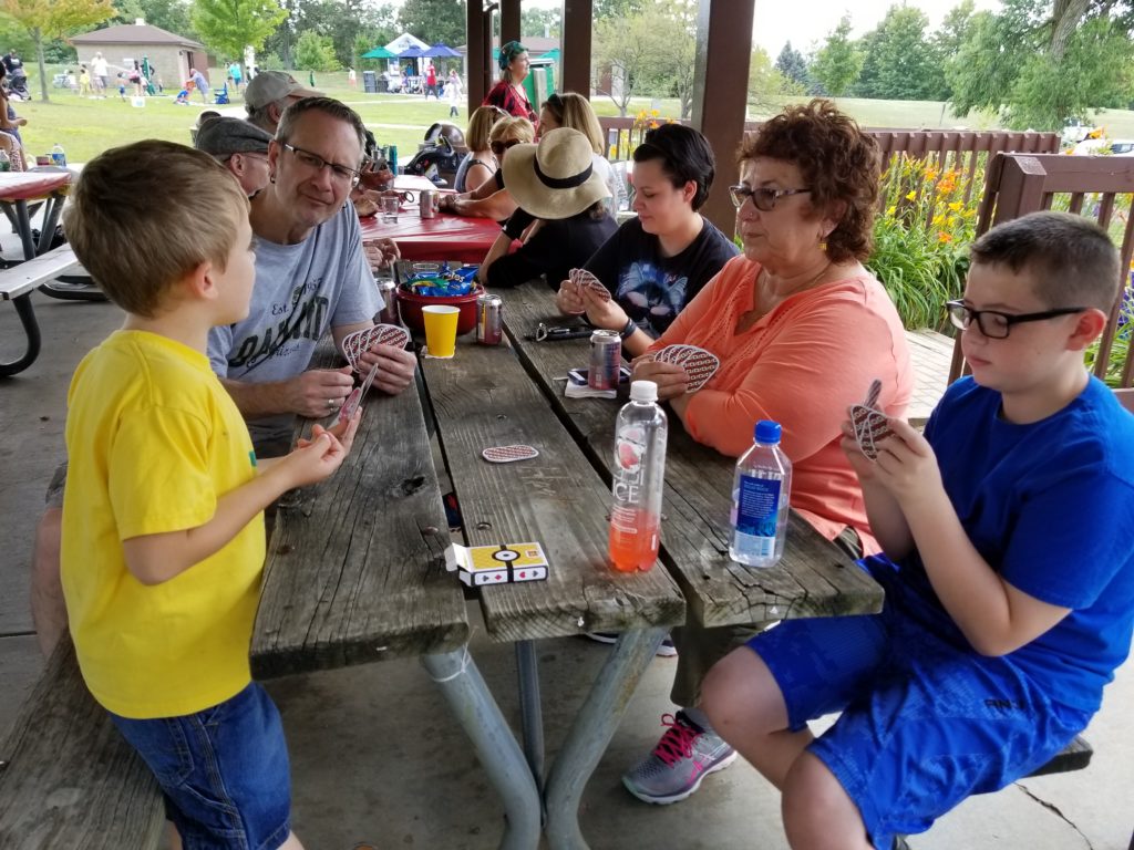 A group of people of various ages sit at a picnic table playing cards together.