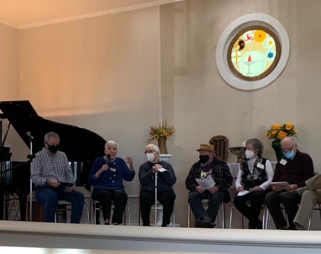 6 UUCF members sit in a circle near the pulpit, sharing historical and personal stories during our Ancestor Service. A piano sits behind the group on the left, and UU chalice behind. A stained class window representing a multitude of religions shines on the wall behind and above the chalice.