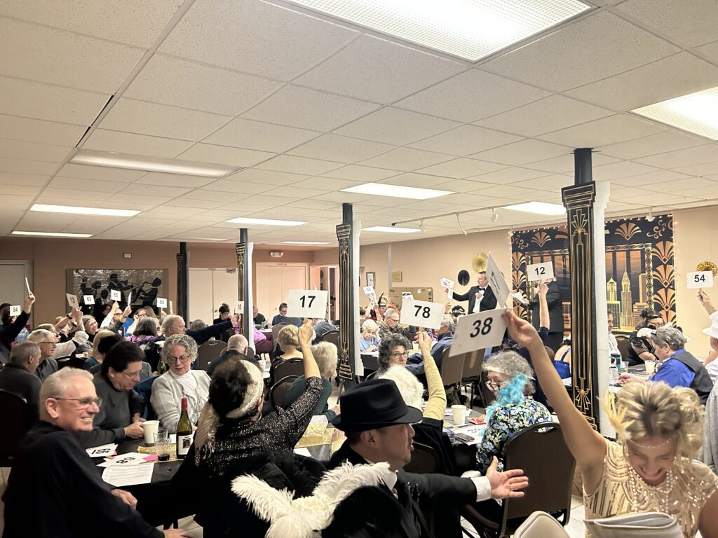 A wide shot of a social hall filled with people sitting at tables with bidding paddles in the air. The people and decorations are black and gold in a 1920s theme.